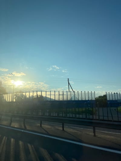 The image depicts a roadside view with a clear blue sky and scattered clouds. The sun is low on the horizon, casting a bright glow and creating lens flare. A street light is visible to the left. In the mid-ground, a dark metal fence runs horizontally, partially obscuring a transparent barrier with vertical supports. Beyond the barrier, trees and vegetation are visible. A utility pole with wires stands in the background. The foreground shows a road with a white line marking the edge. Shadows from the fence and potentially from a vehicle are cast across the road. The scene appears to be located along a highway or major road in the vicinity of Фаніпальскі сельскі Савет, Belarus, given the provided context.