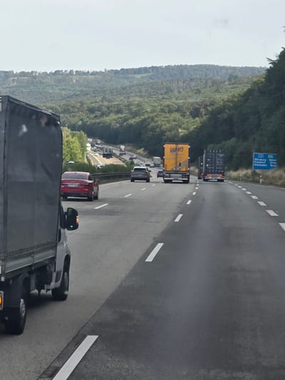 The image depicts a highway scene on a cloudy day, likely in Germany. In the foreground, the rear of a dark-colored truck is visible, obscuring part of the view. Ahead, a red Audi sedan is in the left lane, followed by other vehicles. The right lanes show several trucks, including a yellow one with "DECKO LOGISTIK" branding and a dark container truck. A blue highway sign in the distance indicates distances to Würzburg (158 km), Frankfurt (43 km), and Wiesbaden (18 km). The highway is flanked by dense, green forested hills. The overall impression is one of moderate highway traffic amidst a natural landscape. There is no discernible information to specifically place the scene within Idstein, Germany, although the sign pointing to Frankfurt and Wiesbaden suggests it could be on a route approaching or departing these areas, which are in proximity to Idstein.