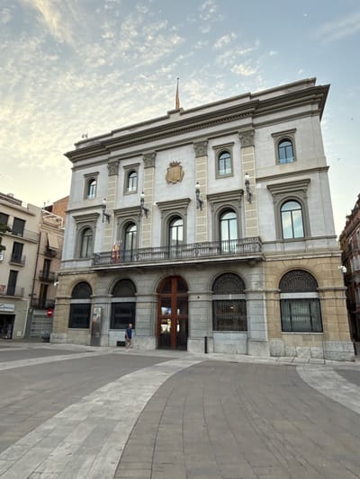 The image shows the Town Hall of Igualada, Spain. It is a large, imposing building with a light-colored facade, featuring ornate architectural details such as columns, arches, and decorative carvings. The building has multiple stories, with several arched windows on the ground floor and upper floors. A balcony with a wrought-iron railing extends across the main facade. Two flags, one appearing to be the Catalan flag, are visible on the balcony. Above the entrance, a coat of arms is mounted. The building is situated in a wide, paved public square. The square has a distinctive pattern of light and dark paving stones, creating curved walkways. A single person, a male wearing a blue shirt and white shorts, is walking across the square towards the building. The sky above is partly cloudy with a few birds visible. Adjacent buildings, including a taller, narrower building with balconies, are visible to the left of the Town Hall. The overall impression is of a civic building in a well-maintained urban setting.