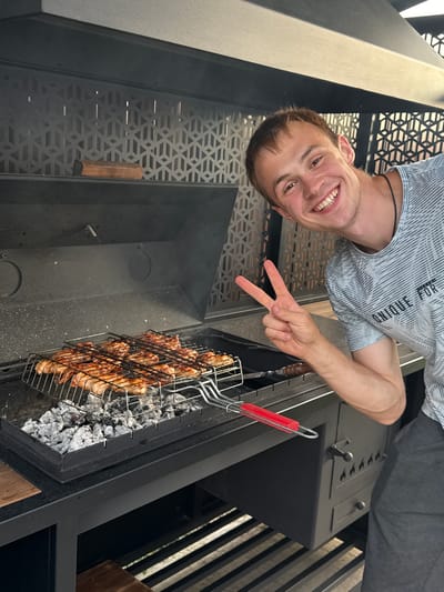 The image shows a young man in Starokorsunskaya, Russia, smiling and making a peace sign while grilling chicken. He is wearing a grey t-shirt with a striped pattern and text that reads "UNIQUE FOR". The chicken is in a grilling basket over hot coals, and smoke is visible. The man is positioned in the foreground, with the grill and a portion of a black outdoor kitchen unit behind him. The overall scene suggests a casual, outdoor cooking activity. The lighting indicates it's daytime.