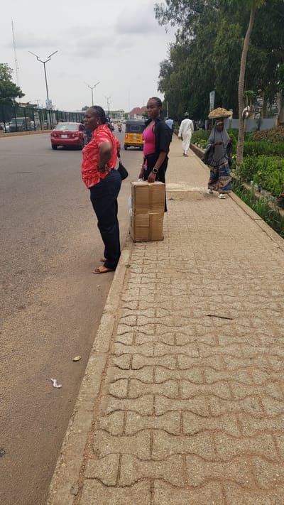 In Unguwar Shanu, Nigeria, on a cloudy day, two women stand by the side of a paved road. The woman on the left, wearing a red patterned shirt and dark jeans, has her hands on her hips. The woman on the right, in a black jacket over a pink top, holds a cardboard box in her hands. Behind them, a person in a white robe walks away, and another woman, wearing a headscarf and a colorful skirt, carries a basket of goods on her head. Traffic, including a red car and a yellow tricycle, is visible on the road. The sidewalk is made of interlocking concrete pavers. Trees and greenery line the far side of the road.