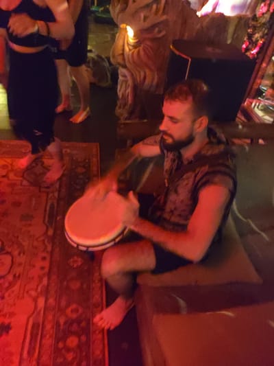 A seated man plays a drum in a dimly lit indoor setting. The man has a beard and wears a patterned sleeveless shirt and dark shorts. He is barefoot and sits on a cushioned surface. He holds a drum with a light-colored skin and dark rim. A patterned rug covers the floor near him. Other individuals stand nearby, and decorative elements such as a sculpted object and a cabinet are visible in the background. The location is Lokaserena, Indonesia.