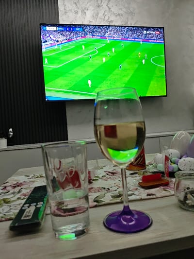 The image captures an indoor domestic scene within the city of Opaka, Bulgaria. A wall-mounted, flat-screen television is prominently displayed, showing a football (soccer) match between teams labeled "RMA" and "MCI," with a score of 0-0 and a game clock indicating 0:49. The wall behind the television features dark, vertically ribbed paneling on the left and a light-colored, textured surface on the right. A light-toned shelf or counter runs horizontally below the television, holding a white decorative bust with a metallic face on its left side.

In the foreground, a light-colored table is partially covered by a floral table runner. Arranged on the table are several items: a clear glass approximately half-filled with clear liquid, a dark-colored television remote control positioned next to it, and a wine glass with a clear bowl containing a pale yellow liquid and a distinctive purple stem and base. Behind the wine glass, a partial view of an orange-brown cylindrical packaged snack is visible. To the right of the wine glass, a small red object, possibly a lighter, is present. Further right, a small clear glass jar containing cigarette butts and ash is visible. On the far right of the table, a collection of white, speckled egg-shaped decorations and smaller pastel-colored candies or stones suggests an Easter theme. No individuals are depicted in the scene, which implies a setting for leisure and entertainment.