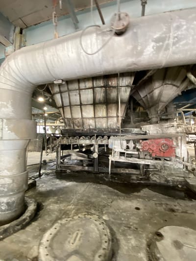 The image depicts an industrial setting within the city of Police, Poland. The scene is dominated by large, insulated pipes and industrial machinery. A prominent feature is a large, gray, insulated pipe curving into the frame from the left, leading towards a complex structure with multiple chutes and a series of rollers. To the right of the rollers, a red gear mechanism is visible, suggesting active machinery. The floor appears wet and dirty, with puddles and debris scattered around. The overall environment suggests a processing plant or factory, likely involved in material handling or a manufacturing process. No people are present in the image.