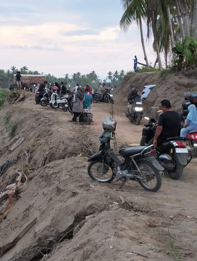 A crowded scene unfolds at what appears to be a coastal location, possibly a tourist destination in Kapa, Indonesia. Numerous individuals and motorcycles are congregated on a dirt road or embankment. The ground is eroded, exposing underlying soil and tree roots.

People: Many people are present. They are diverse in age and appearance, mostly dressed casually. Some are on motorcycles, others are standing and interacting. One man is positioned on an elevated section near palm trees.

Subjects: Motorcycles of varying types and conditions are a central element, indicating a common mode of transportation and congregation.

Objects: Besides motorcycles, other identifiable objects include palm trees and a structure with a thatched roof.

Actions: People are congregating, possibly observing something out of frame, riding motorcycles, or simply enjoying the location.

Interactions: The scene suggests a social gathering. Interactions include people standing together, some on motorcycles interacting with others, and a general sense of collective activity.

Environment: The setting is coastal with palm trees dominating the backdrop. The terrain is uneven with apparent erosion of the ground. The time of day appears to be late afternoon or early evening, indicated by the lighting.