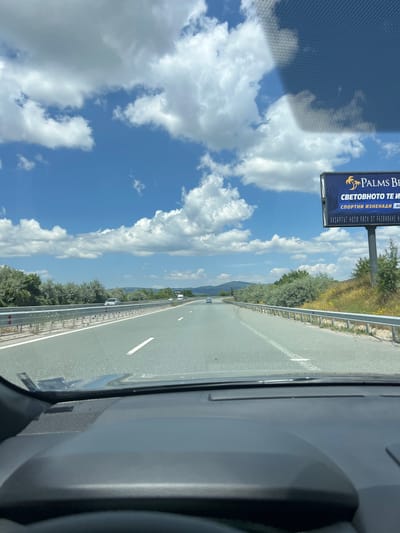 A first-person perspective shot taken from inside a vehicle on a highway. The highway stretches into the distance under a partly cloudy blue sky. Green trees and vegetation line both sides of the road, with mountains visible in the far distance. A large billboard is present on the right side of the highway, advertising "PALMS BE" and text in Bulgarian. Several cars are visible on the highway, some in the distance and one in the left lane in the foreground. The dashboard of the vehicle is visible in the lower portion of the image. The scene appears to be in Studena, Bulgaria.
