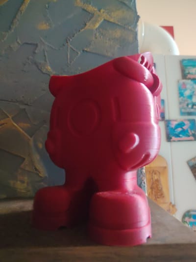 A bright red 3D printed figure resembling a cartoon character is placed on a wooden shelf. The figure is facing to the right and has large, circular eyes and a simple mouth. The background features a textured, blue-gray wall with yellow accents, and several small framed pictures are visible on a white surface behind the figure. A white pipe is also present in the background. The scene is indoors, and the lighting suggests it might be daytime. The image does not provide any information about the city of Сухарево, Russia.
