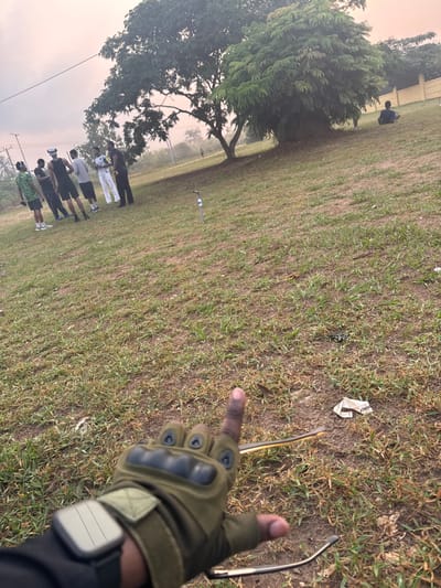 The image captures an outdoor, grassy area in Obinze, Nigeria, under a hazy sky with a pale orange tint. In the immediate foreground, a person's right hand, clad in an olive-green fingerless tactical glove featuring black knuckle protection and a light-colored smartwatch, extends with the index finger pointed upward. A pair of sunglasses is partially visible, draped over or near the hand. The ground is covered with a mix of green and dry brown grass, interspersed with patches of bare earth and scattered debris, including a crumpled white wrapper. A light blue, slender pipe or post protrudes from the ground in the mid-left. In the left background, a group of approximately seven males stands on the grass, casually dressed in various shirts and pants/shorts, appearing to be in conversation or observing their surroundings. In the right background, a large, dense green tree stands prominently. To its right, an individual is seated on the ground near a light-colored wall or fence, partially obscured by the tree. Distant power lines and additional foliage are visible along the horizon.