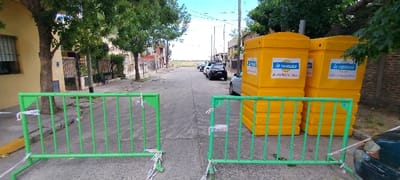 This image captures a residential street scene in San Fernando, Argentina, during the daytime under a partly cloudy sky. The foreground is dominated by two bright green metal barriers, positioned to restrict access to the street. These barriers are secured with white and red warning tape, suggesting a temporary closure or a designated area.

On the right side of the street, behind the barriers, are several large, bright yellow containers, stacked in at least two sets. These are clearly identifiable as portable toilets, rented from a company whose logo "La Rumana" is prominently displayed on their sides. Below the logo, text for the company's website "www.larumana.com.ar" and a phone number, "2323 481-229 / 481-228," are visible. The presence of these rental units and the barriers strongly indicates an ongoing event, construction project, or public works requiring temporary facilities and area demarcation.

The street itself is paved and extends into the distance, where several cars are parked, including a black SUV and a lighter-colored sedan. On both sides of the street, trees with green foliage line the path, and residential buildings are visible. To the left, a light yellow house with a barred window can be seen, while the right side features a brick wall and other structures. Utility poles and overhead wires are visible further down the street. The scene is devoid of people, giving it a quiet, functional atmosphere, set within a typical Argentinian neighborhood.