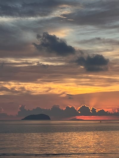 This image captures a serene sunset over the sea near Cuyo, Philippines. A silhouetted island sits on the horizon, its dark shape contrasting with the vibrant sky. The water reflects the warm hues of the sunset, with ripples creating a textured surface. Clouds are scattered across the sky, some dark and dramatic, others illuminated by the setting sun, casting rays of light. A small boat is visible in the distance, adding a sense of scale and tranquility to the vast seascape. The overall scene is peaceful and picturesque, showcasing the natural beauty of the location.