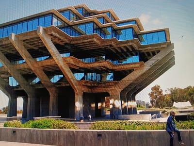 This is a photograph of the Geisel Library at the University of California, San Diego. The Brutalist-style building, constructed primarily of concrete and glass, features a distinctive tiered design with cantilevered floors. Several people are visible in the image: two individuals stand near the entrance on a lower level, and another person is seated on a concrete wall in the foreground, looking to the right. The building is surrounded by landscaping, including green bushes and trees, and a bright sky is visible in the background. The scene depicts an architectural landmark on a clear day.