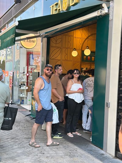 The image captures a street-level view of a "TEA BAR" establishment in Ashdod, Israel. The storefront features a prominent green awning, beneath which a circular, illuminated sign with "TEA BAR" text is visible. The entrance is open, revealing an interior with patterned yellow walls and hanging globe lights. Several people are present outside the establishment.
In the foreground, a man with a beard, wearing sunglasses and a blue tank top with navy blue shorts, stands facing the viewer. He has his right hand in his pocket and is wearing Birkenstock sandals. To his left, a black shopping bag with the word "UAR" is visible on the ground.
Behind the man, a group of people are either entering or exiting the Tea Bar. A woman with dark hair, wearing a white camisole and black pants, is holding a pink phone. To her right, a person in a grey t-shirt with a printed graphic on the back stands in the doorway, wearing light blue jeans. Other individuals are partially visible further inside the establishment.
The ground is paved with cobblestones and grey concrete tiles. The overall scene suggests a casual, urban environment on a bright day.