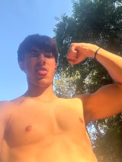 A young male subject, appearing shirtless, is depicted in a low-angle outdoor selfie taken in the city of Marikostinovo, Bulgaria. He has dark hair, styled with bangs, and a light amount of facial hair. The subject is actively winking with his right eye (viewer's left) and extending his tongue in a playful expression. His left arm (viewer's right) is flexed, showcasing a bicep, and he wears a dark, possibly braided, bracelet on his left wrist. The background features a clear, bright blue sky in the upper left, transitioning to dense green foliage of trees in the upper right and behind the subject's arm. Strong natural sunlight illuminates the scene from an elevated position, casting distinct shadows on the subject's neck, chest, and arm.