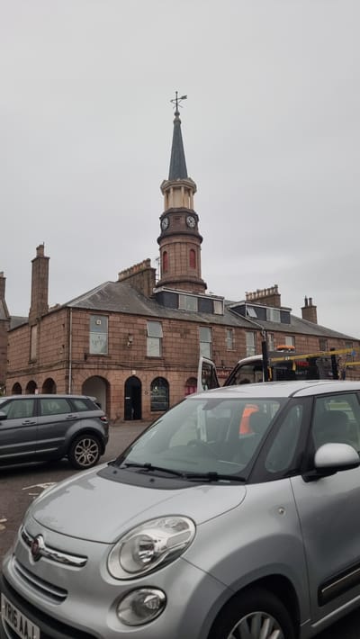 The image shows a street scene in Stonehaven, United Kingdom, featuring a prominent clock tower with a weathervane. The tower is made of stone and has visible clock faces. Adjacent to the tower is a building constructed with reddish-brown stone, featuring multiple windows and arched doorways. A sign on the building reads "The Market".
In the foreground, two cars are parked. The one closer to the viewer is a silver compact car, with its headlights and front grille visible. The other car, parked behind and to the left of the silver car, is darker in color and partially visible.
The sky is overcast, suggesting a cloudy day, possibly with light rain as there are water droplets visible on the silver car. The overall lighting is muted. The scene appears to be a town or city street, likely during daylight hours, given the visibility of the subjects. No people are clearly visible in the image. The atmosphere seems ordinary, with no specific event or activity depicted other than the cars being parked on the street.