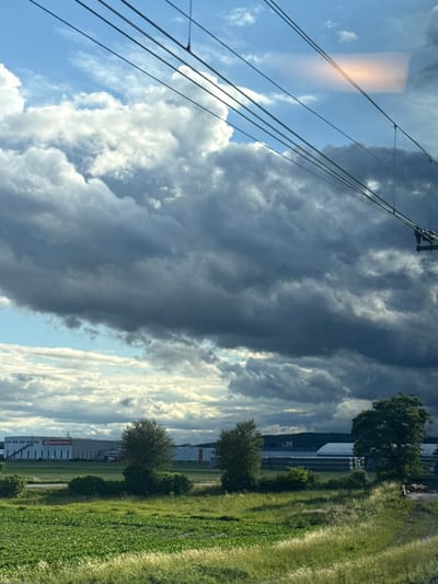 The image depicts a wide landscape view, likely captured from a moving vehicle, given the perspective and the presence of overhead wires. In the foreground, a lush green field of crops stretches across the frame, with taller grasses lining the immediate foreground. Beyond the field, a row of trees, some dense and some more sparse, breaks up the landscape. In the midground, several industrial-looking buildings are visible, characterized by long, rectangular structures with white and red accents. One prominent building features a large red logo. In the background, a dark, hilly horizon is partially obscured by dramatic, dark grey storm clouds that dominate the upper portion of the sky. Bright white clouds are interspersed with the darker ones, suggesting a dynamic weather system. The sky is a mix of blue and grey, with a peculiar, elongated orange-hued light anomaly visible high up, possibly a reflection or atmospheric phenomenon. The overall impression is one of a rural or semi-industrial landscape under an approaching storm. The city of Loosdorf, Austria, is noted as the location, implying the buildings and landscape are situated within or near this Austrian municipality.