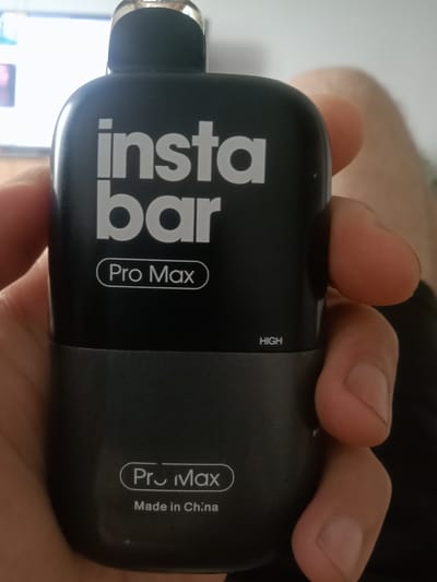 The image is a close-up, indoor, eye-level shot of a person's hand holding a black and gray vape device. The vape device has "insta bar" and "Pro Max" printed on it in white text. The word "HIGH" is also visible in smaller white text. The lower portion of the device is gray and has "Pro Max" and "Made in China" printed on it. The background is blurred, but appears to show a screen or monitor and a hint of a person's leg. The lighting is bright. There is no indication of the location being in the city of Kielce, Poland.