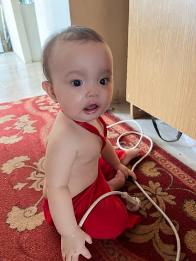 A baby wearing a red garment is lying on a patterned red rug. The baby is facing upwards, with their mouth open and eyes wide. They are positioned next to a wooden surface, possibly a desk or cabinet. A white electrical cord with a plug is visible near the baby's legs and hand, lying on the rug. The scene appears to be indoors, with a carpeted floor and a wooden piece of furniture. The lighting suggests it could be daytime. The baby's expression conveys curiosity or surprise. There is no visible text in the image. The context of Kyzylorda, Kazakhstan is provided.