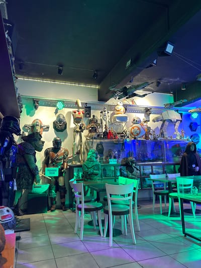 The image shows the interior of a commercial establishment, likely a restaurant or cafe, in Villa General Belgrano, Argentina. The dominant feature is a wall display of Star Wars memorabilia, including action figures, helmets, and model spaceships. The items are arranged in a tall cabinet with glass shelves and are illuminated by blue and green lights. Adjacent to the Star Wars display are tables and chairs, indicating the dining area of the establishment. The tables are lit with green and purple lighting. The overall atmosphere is themed, creating an immersive environment for customers. The wall behind the Star Wars display is dark, contrasting with the brightly lit exhibits. A sign is present, reading "PROHIBIDO TOCAR," meaning "DO NOT TOUCH" in Spanish, indicating the exhibits are off-limits to customers.