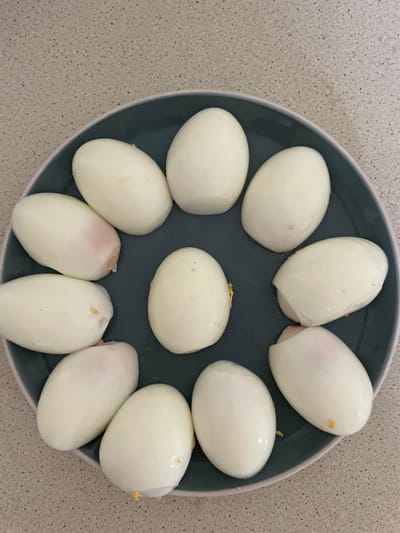 A close-up overhead shot shows a dark gray plate filled with peeled hard-boiled eggs arranged in a circular pattern. The eggs are predominantly white, with some showing slight pinkish tinges or small yellow specks. The plate rests on a speckled light-colored countertop. The image does not contain any information to determine its location within the city of Reading, United Kingdom.