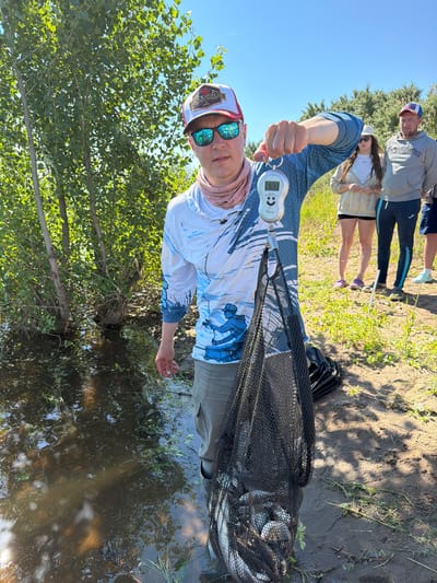 The image depicts a man standing on the bank of a body of water, holding a net filled with fish. He is wearing a long-sleeved fishing shirt, khaki pants, a baseball cap, and sunglasses. A digital scale hangs from his outstretched hand, displaying a weight of 6.33. Behind him, two women and another man are visible, also appearing to be outdoors. The environment suggests a natural setting with trees and vegetation. The location is identified as Tarasovo, Russia.