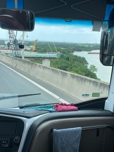 The image shows a view from the driver's perspective inside a vehicle, likely a bus, looking out through the windshield and side mirror. The vehicle is on an elevated roadway, possibly a highway, with a concrete barrier separating the lanes. In the distance, there is a body of water, likely a river, with a long, red barge or ship sailing on it. The landscape is characterized by lush green trees and vegetation on the banks of the river. Construction is evident in the midground, with several large cranes and unfinished bridge structures visible. A billboard with the text "BIC" is also present near the construction site. The sky is overcast with clouds. Inside the vehicle, the dashboard is visible, with a navigation screen and some cleaning cloths draped over the interior. There is a reflection of what appears to be a person's leg wearing maroon-colored clothing in the side mirror. The scene is consistent with construction and development occurring along a waterway in an urban or suburban area of Ho Chi Minh City, Vietnam.