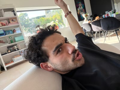 The image depicts a male individual reclining on a light-colored, textured sofa within a residence in Mougins, France. The man has dark, curly hair, a beard, and a mustache, and is looking directly towards the camera with an open-mouthed expression. He wears a black long-sleeved shirt and a brown leather-strapped watch on his raised left wrist.

To the left of the man, a white bookcase holds various objects including books, framed photographs (one displaying an infant), a silver trophy cup, and small boxes. Behind the man, a large window reveals a brightly lit outdoor environment with dense green foliage, trees, and flowering bushes (white and red blooms).

To the right, a dining area is visible, featuring a dark dining table surrounded by dark upholstered chairs with thin metallic legs. The table contains several items: various beverage bottles and cans, a basket filled with what appears to be bread or pastries, a bouquet of red flowers wrapped in white paper, and a black air fryer. On the wall above the dining table hangs a framed artwork featuring a stylized bulldog with a crown, rendered in vibrant, abstract colors. The room has light-colored flooring, and the wall behind the dining table is a contrasting dark color.