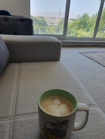 A light-colored textured sofa is in the foreground, with a mug of coffee with latte art on its surface placed on the cushion. The mug has a green rim and a graphic design on its side. In the background, a large window offers a view of trees and distant mountains, suggesting an outdoor setting. The presence of a sofa and window implies an indoor space, likely a living room or lounge. The overall scene is peaceful and relaxed, with the coffee suggesting a moment of leisure. The location of Kheyrabad, Iran, in the context of this image is not visually determinable.