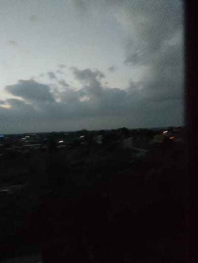 The image presents an elevated, wide-angle view of Juja ward, Kenya, captured during low-light conditions, consistent with dusk or night. The upper portion of the frame is dominated by a heavily clouded sky, displaying variations of dark grey and lighter grey tones. The horizon line is visible beneath the cloud cover. The mid-ground and foreground are significantly underexposed, appearing largely dark, but indistinct silhouettes suggest a landscape populated by trees and structures. Across the distant horizon, a scattering of blurred, artificial light sources is visible, appearing as streaks and points of white and orange illumination, indicative of streetlights, vehicles, or building lights within an urban or semi-urban area. No people are discernible, and no specific actions or interactions are evident. A dark, vertical object, likely a window frame or structural element, partially obstructs the far right edge of the composition.