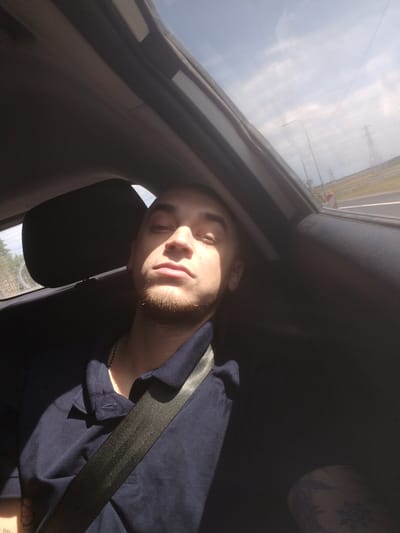 In Cherkasovo, Russia, a young man is captured in a selfie taken from the backseat of a car. Sunlight streams through the car's sunroof, casting harsh shadows across his face and dark blue polo shirt. He wears a silver chain necklace and a black seatbelt diagonally across his chest. The background shows a glimpse of the sky and distant power lines. The interior of the car is dark, with the headrest of the seat in front of him partially visible. The image is shot from a low angle, emphasizing the man's features and the dramatic lighting.