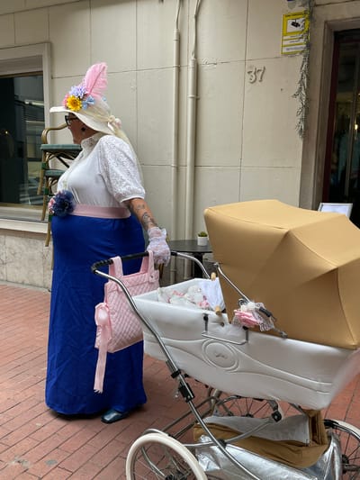 A woman in elaborate period clothing stands next to an ornate baby carriage on a brick walkway. The woman wears a wide-brimmed hat adorned with feathers and flowers, a white lace top, a long blue skirt, and lace gloves. The baby carriage has a white body, a tan umbrella, and silver metal wheels. The number "37" is visible on the building behind the woman, along with a surveillance camera sign. The scene is likely set in Terrassa, Spain.