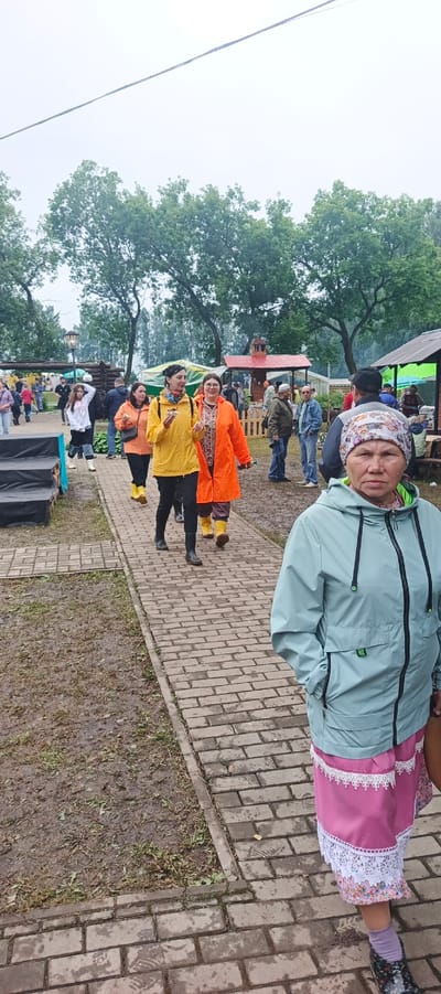 The image depicts an outdoor scene in the village of Staryye Bygi, Russia. A group of people are walking on a brick path, many wearing brightly colored raincoats and boots, suggesting it may be raining or has recently rained, as the ground appears wet and muddy in places. In the foreground, a woman wearing a light blue jacket and a pink skirt with white lace trim is looking towards the camera. Behind her, a woman in a yellow raincoat and another in an orange raincoat are walking, one of them holding food. Several other individuals are visible in the background, along with tents, trees, and some structures that resemble market stalls or pavilions. The overall atmosphere suggests a community event or festival taking place outdoors.