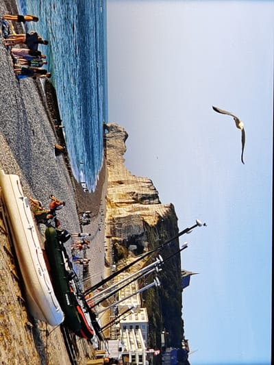 This is a vertical, eye-level shot of a beach in Étretat, France, with a dramatic cliff face in the background. The scene is bathed in bright, clear daylight, suggesting fair weather.
In the foreground, several inflatable boats are lined up on the pebble beach, with one larger, white boat on the left and two smaller, darker ones to its right. These boats appear to be used for recreation or fishing. Scattered along the beach are several people, some standing and others sitting or walking, enjoying the coastal scenery. One group of people is gathered near the water's edge.
The midground features the imposing, layered rock formations of the Étretat cliffs, characteristic of the region. The sea stretches out to the right, appearing calm and blue. In the sky, a single bird, possibly a seagull, is captured mid-flight, soaring gracefully against the pale blue expanse.
Further into the background, buildings can be seen nestled against the base of the cliffs, indicating a small town or settlement. The overall atmosphere is one of a leisurely summer day at the seaside, with a focus on natural beauty and outdoor activities. There is no visible text in the image.