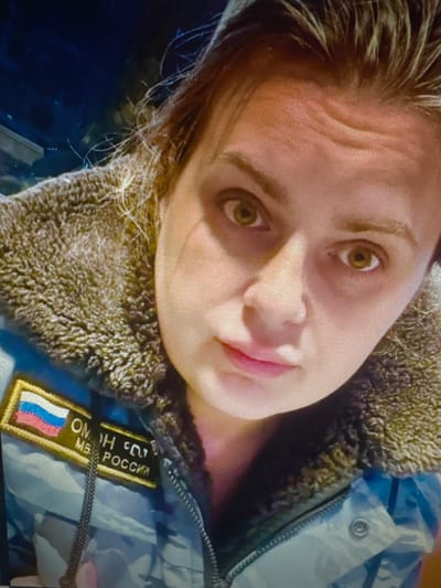 A close-up, waist-up, indoor selfie of a woman with light brown hair and green eyes. She is wearing a camouflage jacket with a fur-lined collar. A patch on the left side of her jacket displays the Russian flag and text that appears to read "ОМОН МВД РОССИИ" which translates to "OMON Ministry of Internal Affairs of Russia". The background is dark, suggesting nighttime or an indoor setting with low lighting. The woman's expression is neutral. The location is specified as Protasovo, Russia.