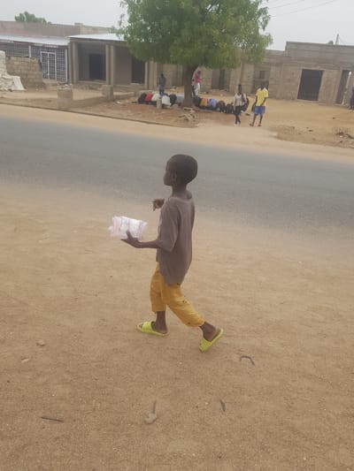 A young person is walking on a dirt shoulder next to a paved road in Dutsinma B, Nigeria. The person is wearing a gray shirt, yellow pants, and green sandals, and holding a plastic bag. On the left side of the road are a partially constructed building and a group of people gathered under a tree.
