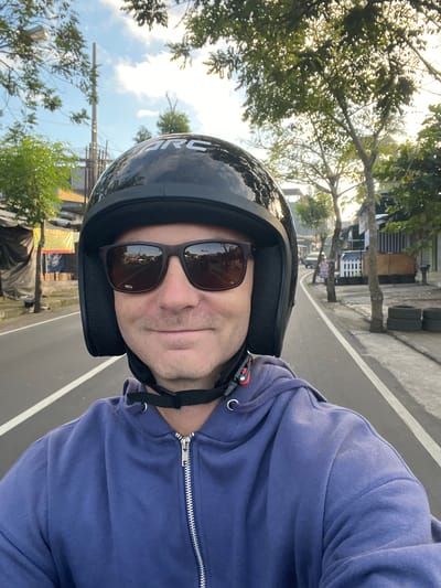 The image shows a man wearing a black helmet and sunglasses, riding a motorcycle on a street in Abiansemal, Indonesia. The man is smiling and looking at the camera. He is wearing a blue hoodie. The street is paved and has white lines marking the lanes. There are trees and buildings on both sides of the street. The sky is blue with some clouds. There are other vehicles visible in the background, including a car and a motorcycle. The scene appears to be during the daytime, with the sun casting shadows.