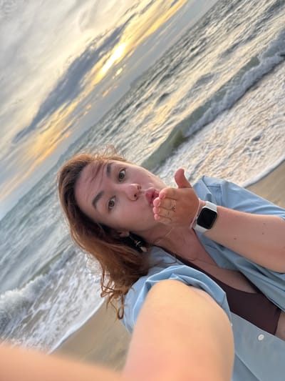 In the city of Tong Yang, Thailand, a woman with auburn hair takes a selfie on a beach at sunset. She is wearing a light blue open shirt over a brown bikini top and has a white smartwatch on her left wrist. The woman is leaning into the camera, making a kissing gesture with her lips pursed and her left hand extended towards the lens. The ocean waves are visible in the background, with the setting sun casting a warm glow on the clouds and the water. The beach sand is also visible in the foreground.