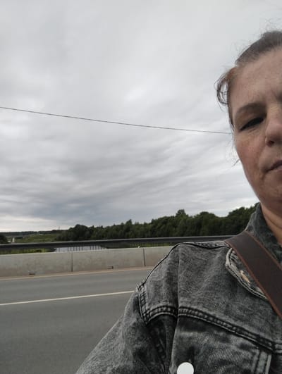The image is a selfie taken outdoors in Kingisepp, Russia, on a cloudy day. The foreground is dominated by the right side of a woman's face and upper body, who is wearing a distressed denim jacket. Her hair is tied up, and stray strands are visible. She has freckles on her face and is looking towards the camera with a neutral expression. A dark brown strap, possibly from a bag, is visible across her shoulder and jacket.

The background depicts a road and a body of water, likely a river, bordered by a dense line of green trees. A bridge or elevated structure is visible in the distance over the water. A concrete barrier separates the road from the area where the photo is taken. A thin wire stretches horizontally across the upper part of the frame. The overall mood of the image is casual and the weather appears overcast. There is no visible text in the image.