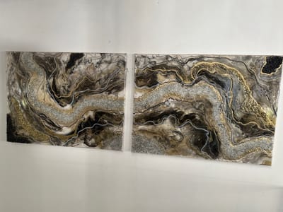 Two abstract art panels, arranged horizontally as a diptych, are mounted on a plain white wall in Marcoussis, France. Each panel presents a glossy, resin-based composition featuring swirling, geode-like patterns. The color palette primarily consists of black, dark brown, gold, silver, and white, forming undulating layers and inclusions. Integrated within the resin are textural elements of crushed silver and gold glass or glitter, which create prominent shimmering bands. Fine, irregular lines in black, gold, and light blue delineate the internal contours and separations within the artwork. Areas of darker, coarser granular material are visible, specifically in the upper-right quadrant of the right panel and the lower-left quadrant of the left panel. The two panels are positioned with a narrow vertical separation between them. The background is a stark white wall exhibiting minor surface imperfections. No individuals, active subjects, or interactions are present in the scene.