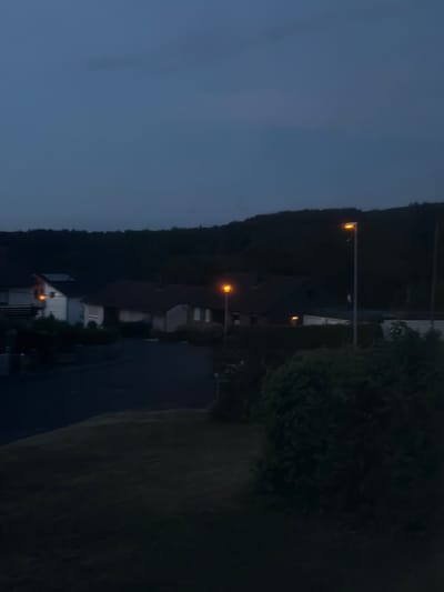 The image depicts a residential area in Idar-Oberstein, Germany, during dusk or early evening. The sky is a dark, overcast blue, indicating low light conditions. In the mid-ground, several houses are visible, characterized by their pitched roofs and illuminated windows, suggesting occupancy. One house on the left has a light visible through a window and a solar panel on its roof. Streetlights with orange illumination are present, one in the mid-ground near the houses, and another further to the right, casting light onto the asphalt road. The road curves through the scene, bordered by dark green foliage and manicured grass in the foreground. A dense, dark treeline forms the background, suggesting a rural or semi-rural setting. No people are visible in the image. The overall impression is one of a quiet, suburban environment at twilight.