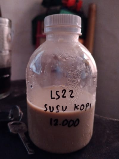 A close-up, low-angle shot focuses on a clear plastic bottle containing a light brown liquid, possibly coffee with milk, condensation droplets covering the exterior of the bottle. Handwritten text in black marker on the bottle reads "LS22" at the top, followed by "Susu Kopi" below, and "12.000" at the bottom. To the left of the bottle, a key with a silver-colored ring is partially visible, resting on a dark, textured surface. In the background, a glass with dark liquid is blurred on the left, and other out-of-focus objects, including what appears to be a wooden pole and some red and green items, are present. The scene suggests a casual, possibly domestic, setting in Cigadung, Indonesia.