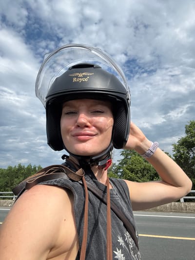 A person wearing a black Royce helmet with a clear visor is taking a selfie outdoors. The person has fair skin, blonde hair, and is smiling. They are wearing a dark gray t-shirt with a graphic print and a brown leather strap across their shoulder. A light purple smartwatch is visible on their right wrist. The background consists of a cloudy sky, green trees, and a road with a white and yellow line. The image is taken from a low angle, looking up at the person. The location is implied to be Suối Tân, Vietnam, based on the surrounding lush vegetation typical of the region and the presence of a paved road.