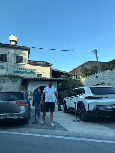 In Jošice, Montenegro, a street-level, full-shot image captures a sunny day outside a restaurant. The facade of the restaurant is made of light-colored stone, with wooden shutters on the upper windows and a turquoise wavy sign spelling out "restaurant" above the entrance. In front of the restaurant, two cars are parked: a silver station wagon on the left, with the license plate "PG LN059", and a white SUV on the right, with the license plate "PG MP 21". A man in a white t-shirt and black shorts, wearing white clogs, walks towards the right of the frame. Behind him, another man in a blue t-shirt and jeans walks towards the restaurant entrance. The ground is paved with light-colored stones, and a street runs in the foreground. The sky is clear and blue, with a power line visible stretched across the upper portion of the image.
