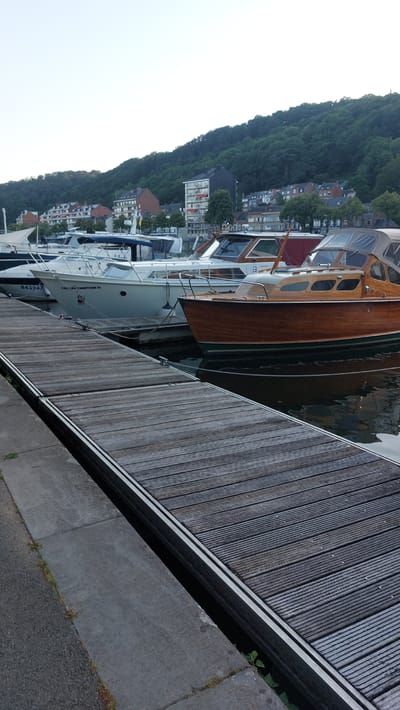 This image captures a waterfront scene in Namur, Belgium. The primary subjects are several boats docked alongside a wooden pier. The boats appear to be of varying sizes and styles, with one prominently featuring a rich, polished wooden hull and cabin. Another boat, a lighter colored motor yacht, has visible text on its side: "COUCOU CHARTEUR III" and "B43247". The boats are moored side-by-side, suggesting they are part of a marina or harbor.

The setting is a waterfront, with a town or village nestled on a hillside in the background. The hillside is densely covered with trees, and the buildings of the town are visible through the foliage, indicating a somewhat urbanized area integrated with nature. The water is dark, reflecting the overcast sky.

The time of day appears to be late afternoon or early evening, given the diffused light and the general atmosphere of the scene, which lacks harsh shadows. The weather seems to be cloudy or overcast, contributing to the muted colors and soft lighting. There are no people visible in the image, focusing the attention on the vessels and the environment. The overall mood is tranquil and still, emphasizing the quiet ambiance of the docked boats. The concrete embankment in the foreground, along with the wooden planks of the pier, provide a textural contrast to the organic shapes of the boats and the hillside.