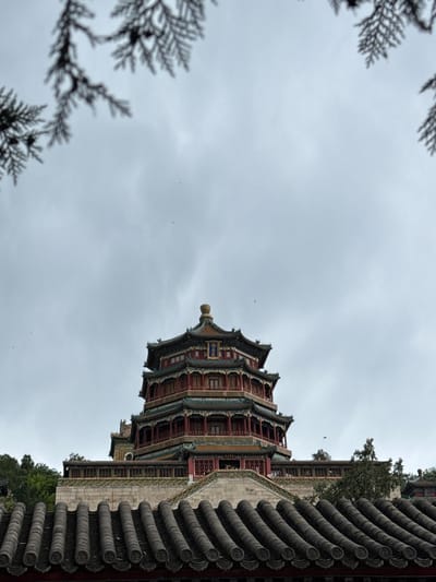 The image displays a towering, multi-tiered Chinese pagoda-style structure, specifically the Tower of Buddhist Incense (Fo Xiang Ge) at the Summer Palace, located in the Haidian District, Beijing, China. The structure is predominantly red and green with ornate detailing, featuring multiple eaves and a golden finial at its apex. It rises from an elevated stone platform, revealing other traditional buildings and lush green trees around its base. In the immediate foreground at the bottom, a row of dark grey traditional Chinese roof tiles, each with a decorative circular end, partially frames the lower part of the scene, indicating another structure closer to the viewer. The upper portion of the image, including the top of the pagoda, is set against a uniformly overcast, light grey sky, with blurred dark green coniferous foliage framing the top left and top right corners. Several tiny dark specks, likely birds, are visible against the sky. No individuals are present in the frame.