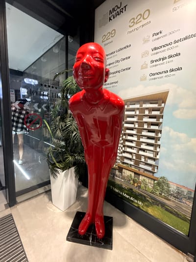 A red, glossy, humanoid sculpture with a child-like, smiling face stands in the foreground. It is positioned on a black marble base. Behind the sculpture, a large poster is displayed, featuring an illustration of a modern apartment building and text in Serbian, indicating it is an advertisement for a residential complex named "MOJ KVART" in Ilidža, Bosnia and Herzegovina. The poster lists various amenities and their distances, such as "Parking mjesta" (parking spaces), "Park" (park), "Vilsonovo šetalište" (Vilson's promenade), "Srednja škola" (high school), and "Osnovna škola" (elementary school). To the left of the sculpture and poster, a glass-fronted building is visible, reflecting light and showing a glimpse of a hotel interior with chandeliers and a woman wearing a plaid shirt and shorts. A potted plant with large green leaves is situated between the sculpture and the poster. The floor is tiled and a metal grate is visible in the lower left corner.