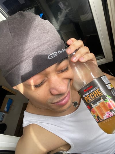 In Plaisir, France, a young adult male is depicted indoors, positioned near a window. He wears a dark gray Kalenji beanie and a white ribbed tank top, with a silver-colored chain necklace and a circular earring in his left ear. His eyes are closed, and he has a subtle smile. Visible freckles are present on his face, along with short facial stubble. He holds a 1-liter clear plastic bottle of Auchan "Instant Gourmand Pêche" (Peach) flavored drink, labeled with "Teneur en fruits 55% minimum," with his right hand near his head. The bottle contains an orange-colored liquid. Strong direct sunlight illuminates the scene, casting distinct shadows across his face and beanie. The background features a white window frame, with dark reflections visible through the glass. A partial view of a shelf or surface with a cardboard box and a blue object is visible to the left.