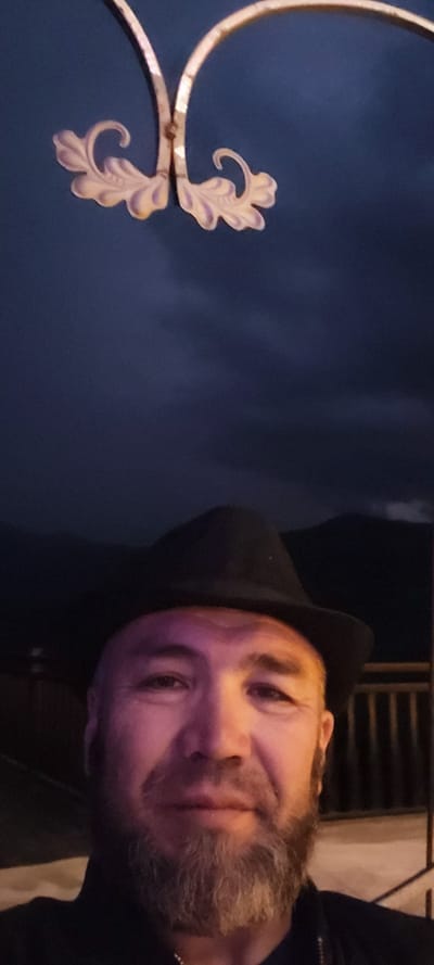 The image is a close-up, vertical, selfie-style photograph taken outdoors at dusk. A man with a beard and a black hat is centered in the lower half of the frame. He is looking directly at the camera, and his face is illuminated by a purplish light. Behind him, a dark, cloudy sky is visible. In the upper portion of the image, a decorative element, possibly part of a lamp or a fence, with ornate, leaf-like shapes, is visible against the dark sky. The background also shows what appears to be a railing and possibly distant hills or mountains. The scene suggests an outdoor setting, possibly a balcony or terrace, in Milisi, Georgia, given the context provided.