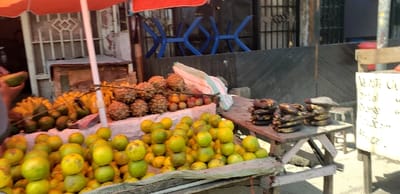The image shows a street vendor selling fruit and other items at a market in Rubavu, Rwanda, near the coordinates -1.668 latitude and 29.268 longitude. The vendor has arranged several fruits for sale, including green avocados, plantains, pineapples, mangoes, and lemons, laid out on a stand covered with a woven cloth. The stand is positioned directly in front of a wooden structure with a closed metal gate. A blue, metal, foldable chair sits on the left of the stand, and a sign is visible in the background, likely advertising prices. A second stand behind the fruit stand displays stacks of dried bananas, presumably for sale. A person is partially visible to the left of the frame, holding a large green avocado. The scene is bathed in natural light, likely midday, and the overall environment is one of a busy, outdoor marketplace.