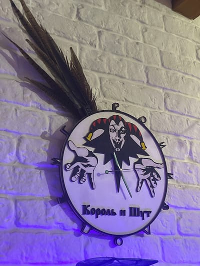The image shows a novelty clock featuring a jester's face and two hands, affixed to a white brick wall. The clock is decorated with dark brown feathers that extend upwards from the top left of the clock face. The text "Король и Шут" (King and Jester) is visible on the clock. The wall is illuminated with purple light from below. The scene is consistent with the interior of the establishment "Korol i Shut" located at 15, Karla Marksa Street, Mineralnye Vody, Russia.