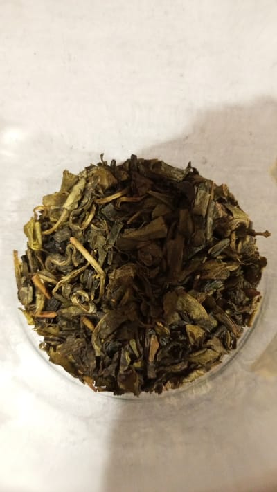 The image shows a pile of loose-leaf tea inside a clear container, likely a jar or glass. The tea leaves are a mix of dark green and brownish-yellow colors, with visible stems and curled leaves. The container is placed against a light-colored, possibly white, surface. The scene appears to be indoors under natural lighting. There is no evidence to indicate that the image was taken in Юдино, Russia. No people are visible in the image.